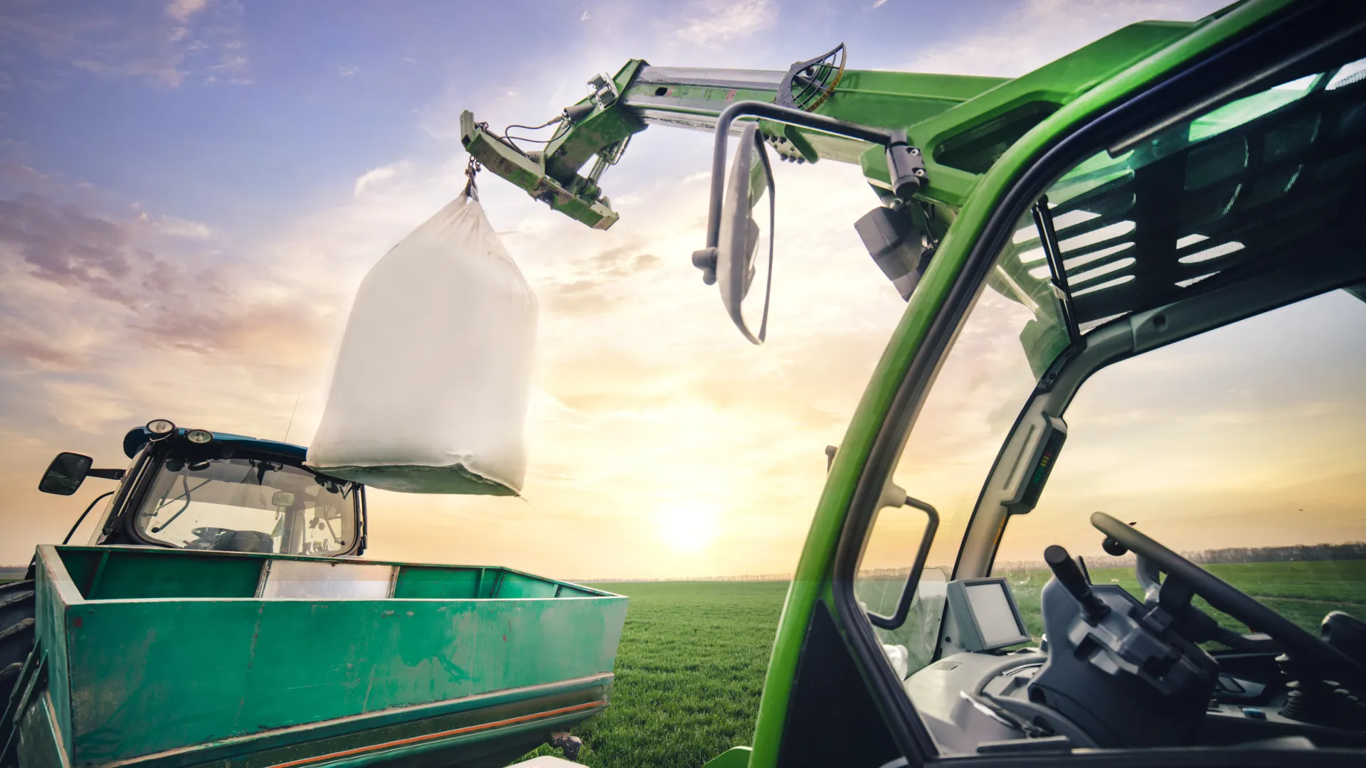 Ein Kranarm lädt im Frühling auf einem Feld einen Sack Dünger auf den Anhänger eines Traktors